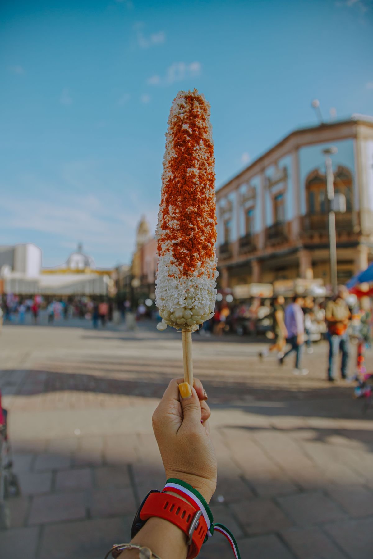 Representative photo of a tall elote on a stick coated in cotija and chile, held up on a plaza